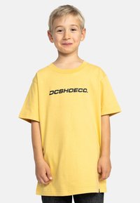 Camiseta de algodón amarilla de manga corta y cuello redondo, con un logo negro "DCSHOECO." en el pecho. Tejido texturizado.