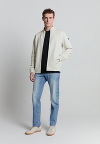 Lichtgekleurde bomberjack met gequiltde textuur, zwart T-shirt, blauwe jeans en beige sneakers. Het jack heeft een staande kraag en een ritssluiting.