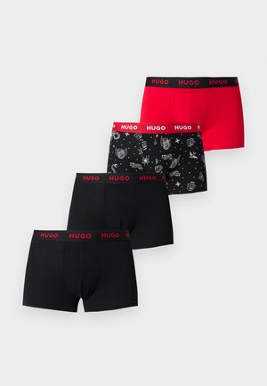 Młody mężczyzna stoi na boso na dywanie, ubrany w czarne bokserki z czerwoną gumą z logo "HUGO", na jednolitym jasnym tle.