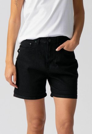 Woman wearing black denim shorts and a white sleeveless top, with one hand in pocket and a flower tattoo on upper arm.