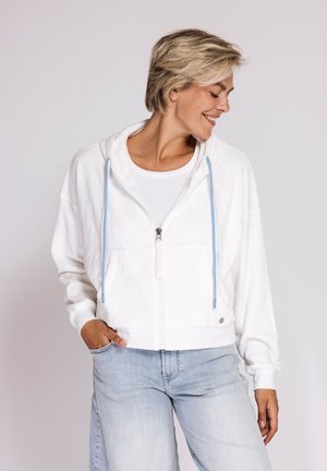 Woman with short blonde hair wearing a white zip-up hoodie with blue drawstrings and light blue jeans, smiling and looking down to the side.
