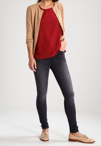 Femme portant un cardigan beige par-dessus un blouson rouge, un jean skinny foncé et des mocassins beige à pampilles, se tenant devant un fond clair uni.