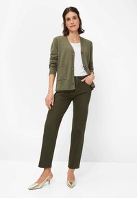 Olivgrüner Cardigan mit V-Ausschnitt, zwei Fronttaschen und Knöpfen, kombiniert mit passenden olivgrünen Hosen und spitzen metallic goldenen Schuhen.