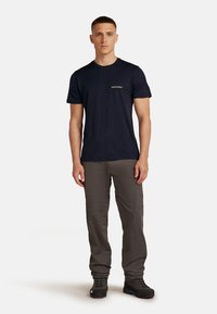 Marineblauw T-shirt met korte mouwen en een ronde hals, voorzien van subtiele groene branding. Draagt bij grijze broek en zwarte wandelschoenen.