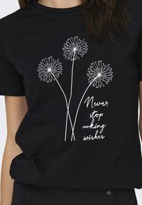 Svart T-skjorte med et hvitt dandelion-blomst grafikk og teksten "Aldri slutte å gjøre ønsker" under, med en avslappet passform og bomullsmateriale.