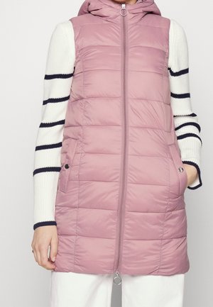 Vrouw die een mouwloze, gewatteerde mauve bodywarmer draagt met een rits en zakken aan de voorkant, over een witte trui met zwarte horizontale strepen.