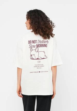 Femme aux cheveux bouclés portant un t-shirt blanc oversized affichant une illustration d'un chien endormi et un texte sur une matinée tranquille.
