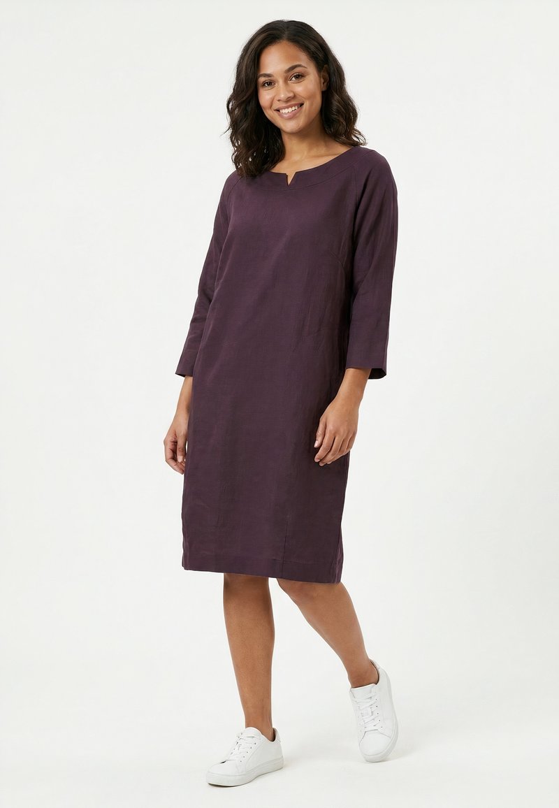 Femme en robe violette longue aux genoux, manches longues, et baskets blanches, debout dans une posture détendue et souriante sur un fond blanc.