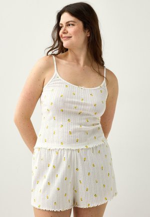 Kvinde med langt brunt hår iført hvid ærmeløs top og shorts med små gule blomster, stående foran en ensfarvet baggrund.