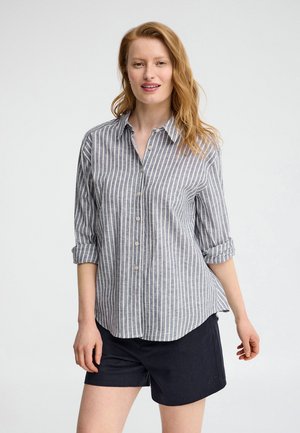 Frau mit heller Haut und roten Haaren trägt ein grau-weiß vertikal gestreiftes, geknöpftes Hemd mit hochgekrempelten Ärmeln und schwarze Shorts vor einem schlichten Hintergrund.