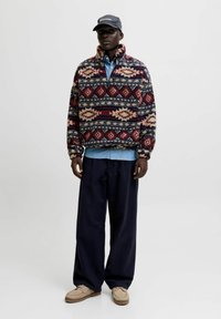 Sudadera de forro polar multicolor con un cuello alto, que presenta patrones geométricos en rojo, azul y crema. Combinada con una camisa de denim y pantalones oscuros.