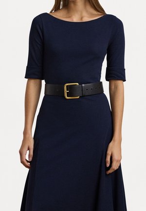 Femme portant une robe bleu marine à manches trois-quarts et une large ceinture noire avec une grande boucle dorée à la taille.