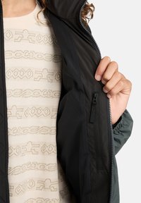 Persona che tiene aperto un giubbotto nero per mostrare la tasca con zip interna e indossa un maglione beige a motivo sottostante.
