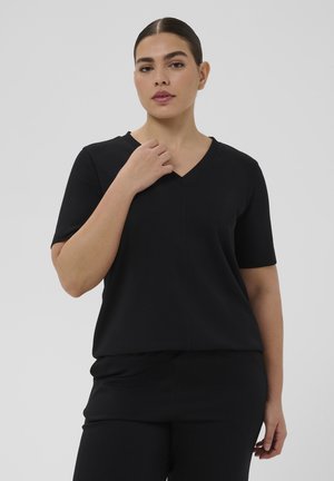 Kobieta o ciemnych włosach związanych w gładki kok, ubrana w czarną bluzkę z krótkim rękawem i dekoltem w serek oraz czarne spodnie, na jednolitym szarym tle.