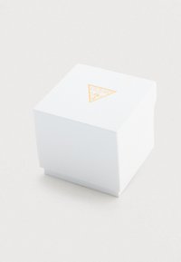 Λευκό κυλινδρικό κουτί με χρυσό λογότυπο Guess που απεικονίζει ένα ερωτηματικό στην κορυφή, τοποθετημένο σε ανοιχτό φόντο.