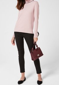 Helles rosa Rollkragensweatshirt mit schwarzem gewelltem Saum, schwarze Skinny Jeans, schwarze flache Schuhe und eine bordeauxrote Handtasche mit silbernem Beschlag.