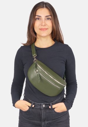 Frau mit braunen Haaren trägt ein dunkles Langarmshirt und schwarze Jeans, sie hat eine grüne Leder-Umhängetasche mit zwei silbernen Reißverschlüssen quer über der Brust.