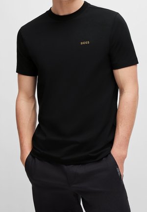 Homme portant un t-shirt noir uni à manches courtes avec un petit logo doré "BOSS" sur le côté gauche de la poitrine, les mains dans les poches d’un pantalon sombre.