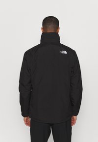 Homme de dos portant une veste noire North Face avec un col montant sur un fond gris clair uni.