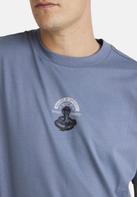 Camiseta de algodón de color azul claro con mangas cortas, que presenta un gráfico de una serpiente negra y el texto "BALANCE DE OPUESTOS INDUSTRIA NATURAL" en blanco.