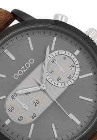 Montre noire avec un cadran gris, indicateurs en argent et deux sous-cadrans. Les caractéristiques incluent un bracelet texturé marron et des accents en acier inoxydable.
