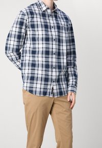 Mężczyzna w granatowo-białej koszuli w kratę na guziki i beżowych spodniach stoi na jednolitym jasnym tle.