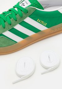 Zelené semišové a syntetické tenisky s bílými pruhy, s nápisem "GAZELLE" ve zlatě a dvěma bílými tkaničkami na světlém pozadí.
