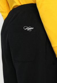 Pantaloni negri cu buzunarul din spate, pe care este brodat logo-ul alb "Kappa", asortați cu un tricou galben cu mâneci lungi.
