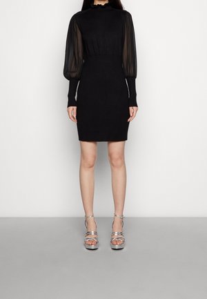 Schwarzes Kleid mit tailliertem Oberteil, hohem, gerafftem Kragen und transparenten Puffärmeln. Kombiniert mit silbernen Riemchenabsätzen. Insgesamt glatte Textur.