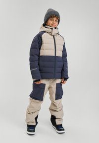 Padded jacket w beżowych i granatowych sekcjach, z zapięciem na zamek i kapturem; zestawiona z beżowymi i granatowymi spodniami narciarskimi z dużymi kieszeniami.