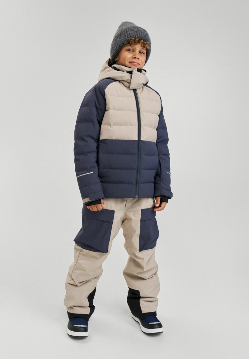 Padded jacket w beżowych i granatowych sekcjach, z zapięciem na zamek i kapturem; zestawiona z beżowymi i granatowymi spodniami narciarskimi z dużymi kieszeniami.