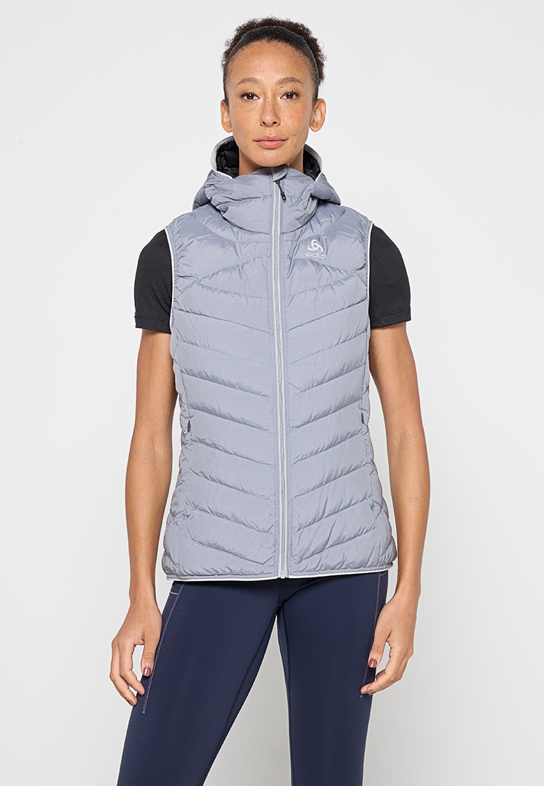 Gepolsterte graue Weste mit hohem Kragen und Reißverschluss vorne, ausgestattet mit einer Kapuze und einem Zickzack-Steppmuster. Getragen über einem schwarzen Hemd.