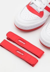 Le sneaker Adidas presentano una tomaia in pelle bianca con accenti rossi, perforazioni e due cinturini testurizzati rossi con dettagli del logo.