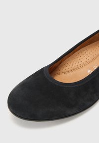 Ballerine en daim noir avec bout arrondi et semelle intérieure perforée et rembourrée couleur caramel sur fond gris clair.