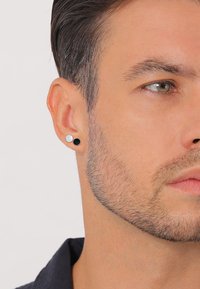 Orecchini circolari in bianco e nero con design a bottone indossati all'orecchio di un modello con capelli corti e barba incolta, su uno sfondo neutro.
