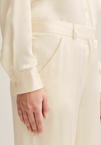 Lähikuva henkilöstä, joka pukeutunut kermaisenvärisiin räätälöityihin housuihin, joissa sivutaskut ja samalla vyöllä, yhdistettynä pitkähihainen napillinen paitaan.