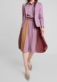 Lawendowa marynarka na fioletowej, falbaniastej bluzce, w połączeniu z plisowaną spódnicą w różowe, pomarańczowe i bordowe kolory. Całość dopełniają buty w motyw skóry węża.