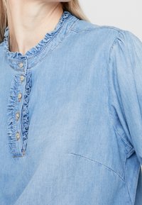 Vaaleansininen farkkupaita, jossa on frillikaulus ja etupaneeli, jossa on kuusi koristeellista nappia ja kerroksittain olevat hihat. Sujuva pinta.