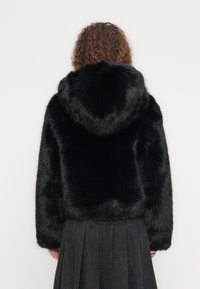 Γυναίκα με σγουρά μαλλιά, ντυμένη με μαύρο μπουφάν με κουκούλα από ψεύτικη γούνα και σκούρα πλισέ φούστα, γυρισμένη ανάποδα μπροστά σε λευκό φόντο.