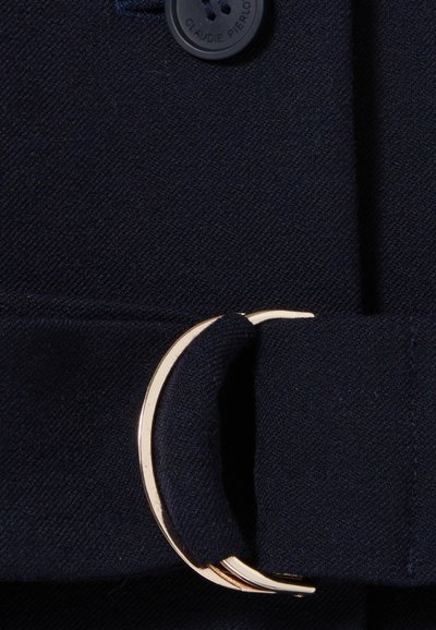 Gros plan sur un tissu bleu marine mettant en avant une boucle en métal doré circulaire et un bouton noir avec le nom de marque embossé, mettant en valeur la texture lisse et les détails de design.