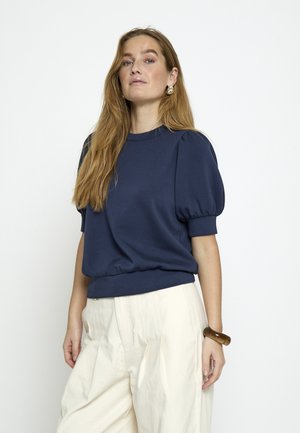 Žena s dugom plavom kosom, odjevena u tamnoplavu bluzu s napuhanih rukava, kremaste široke hlače, zlatne naušnice i smeđu narukvicu, stoji nasuprot bijele pozadine.