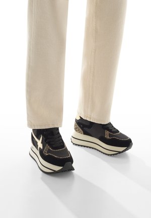 Baskets compensées noires et beige avec des accents de imprimé léopard portées avec un pantalon beige clair sur un fond blanc.