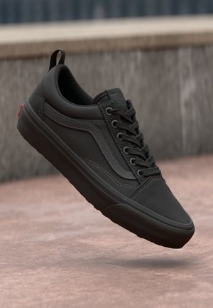 Zwarte lage sneaker met vetersluiting en rubberen zool zwevend boven een geplaveid oppervlak tegen een grijze stenen muur.