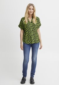 Lühikeste kä sleeve'idega must blouse, millel on rohelised ringikujulised mustrid, koos siniste kitsaste teksaste ja mustade kingadega. Lihtne krae ja mugav lõige.