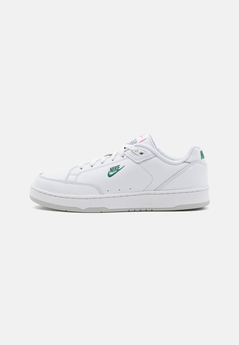 Fehér bőr sportcipők sima textúrával, zöld Nike logóval, szürke talppal és minimalista dizájn elemekkel.
