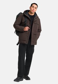 Braune isolierte Jacke mit Kapuze, zwei vorderen Taschen, getragen über einem schwarzen Hemd und einer schwarzen Hose, kombiniert mit schwarzen Turnschuhen und einem dunklen Rucksack.