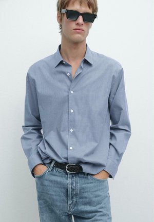 Junger Mann mit schwarzer Sonnenbrille, einem blau karierten Hemd mit Knöpfen und den Händen in den Taschen sowie hellblauen Jeans vor einem schlichten weißen Hintergrund.