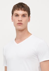 T-shirt blanc à col en V en coton lisse, au design ajusté avec des manches courtes, présentant un look propre et sans couture ainsi que des détails subtils au niveau du col.