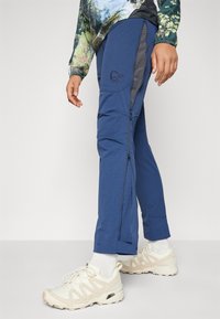 Plave vanjske hlače s bočnim patentnim zatvaračima, teksturiranom tkaninom i tamnosivim naglaskom. Nosi se s bež tenisicama i bijelim čarapama.