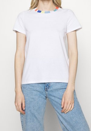 Kobieta ubrana w gładki, biały t-shirt z krótkim rękawem, z wielokolorowym paskowanym wykończeniem pod szyją oraz jasnoniebieskie dżinsy, stojąca na białym tle.